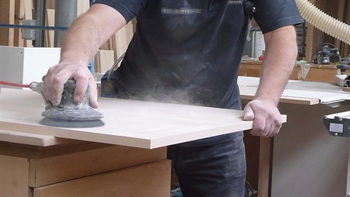 Image of a man using a hand-held power sander on a wooden board, creating airborne dust
